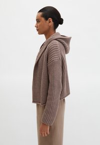 Bruine gebreide cardigan met capuchon, met een geribbelde textuur en een open voorkant, gedragen over een neutraal gekleurde outfit.