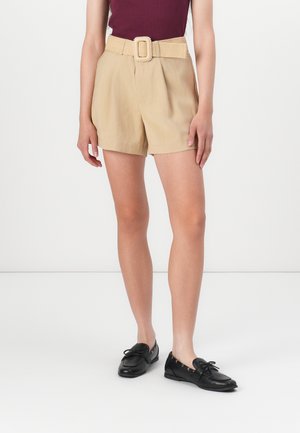 Pantalones cortos beige de talle alto con un cinturón de tejido texturizado que presenta una hebilla cuadrada. Combinados con zapatos de estilo mocasín de cuero negro.