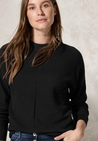Schwarzer strukturierter Strickpullover mit rundem Halsausschnitt und langen Ärmeln, getragen zu blauen Jeans aus Denim mit Knopfverschluss.