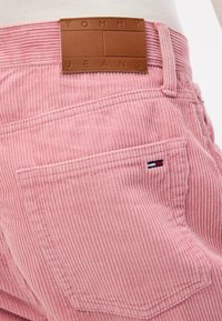 Rožinės velveto kelnės su ruda oda pažymėta etiketė "TOMMY JEANS" ir mažu išsiuvinėtu trijų spalvų logotipu ant galinės kišenės.