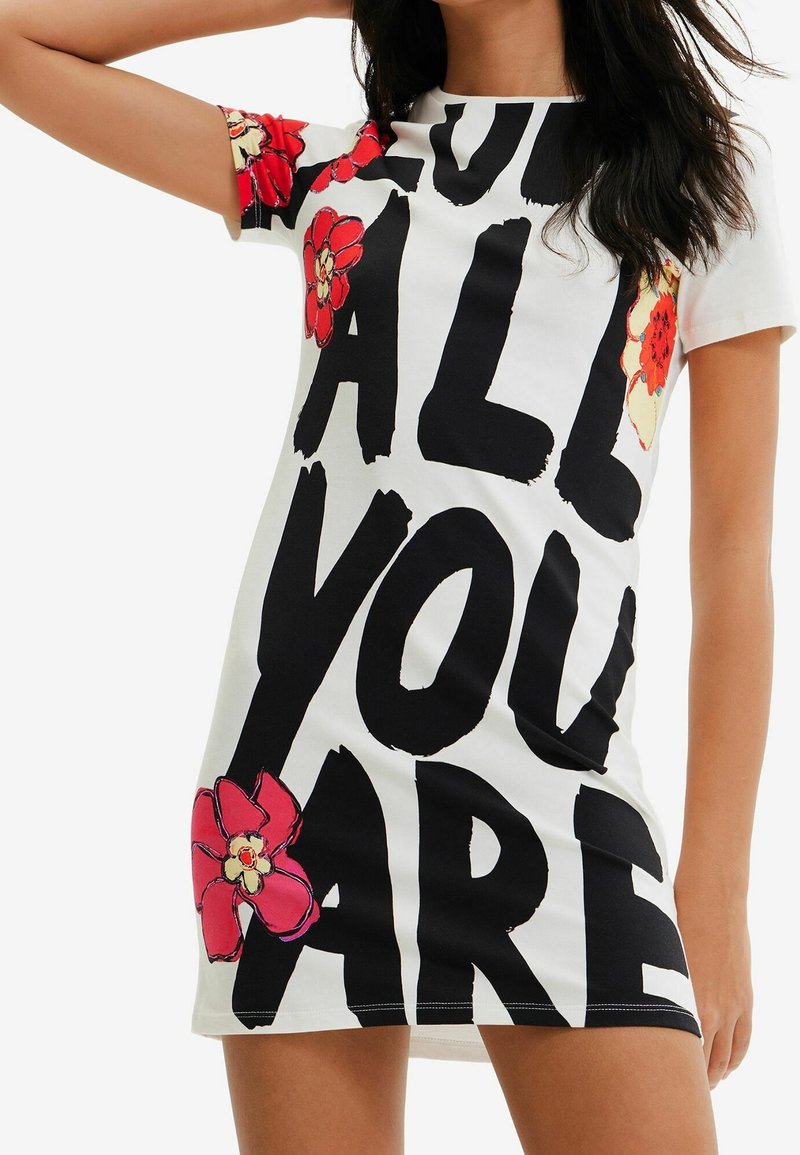 Valkoinen lyhyt mekko, jossa on rohkeaa mustaa tekstiä "LOVE ALL YOU ARE" ja punaisia kukka-akselteja hihoissa, valmistettu sileästä ja pehmeästä kankaasta.