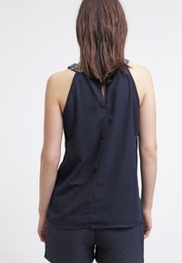 Marinblå ärmlös topp med djup V-back och texturerade dekorationer på axelbanden. Slät tygkvalitet, avslappnad passform.