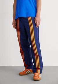 Mørkeblå bukser med et mønster over hele, der har orange og røde striber langs siderne. Parret med orange sneakers og en blå skjorte.