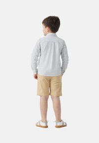 Bambino che indossa una maglietta a maniche lunghe azzurro chiaro e bianca a righe, pantaloni corti color sabbia e sandali bianchi con dettagli grigi, visto da dietro.
