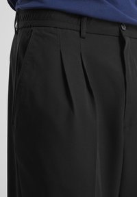 Close-up of black trousers with pleats, side pockets, belt loops, elastic waistband, and a buttoned fly, worn with a blue top.