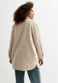 Chaqueta de forro polar beige con un collar alto, hombros caídos y un ajuste ligeramente sobredimensionado. Presenta aberturas laterales y mangas largas.