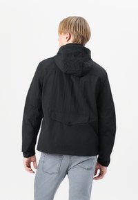 Black jacket made of lightweight material, featuring a hood, a back pocket with a flap, and fitted cuffs. Worn with light gray jeans.
