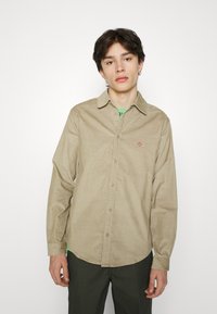 Camisa de pana beige con mangas largas, cierre de botones en el frente, y detalle de logo bordado en el pecho, usada sobre una camiseta verde.