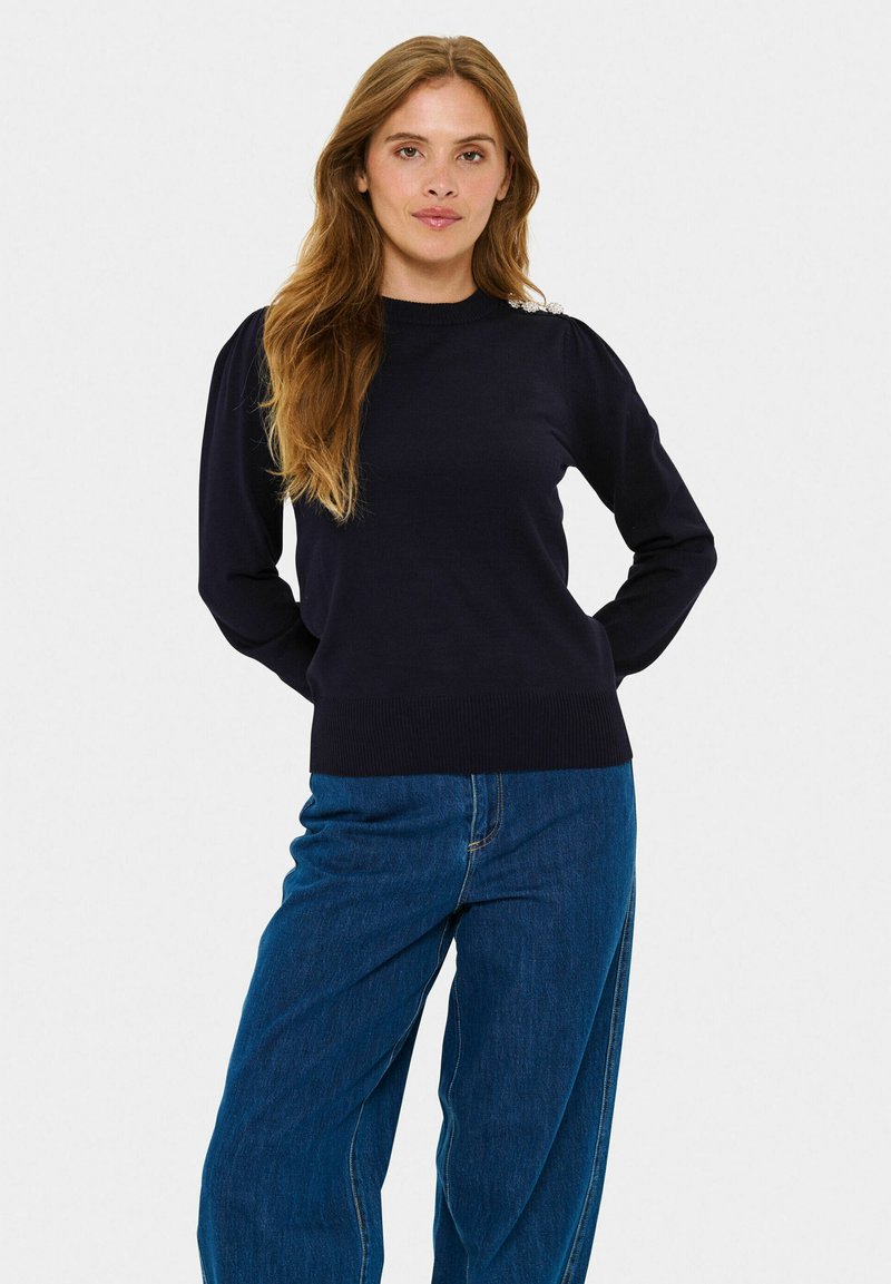 Granatowy sweter wełniany z długimi rękawami i ściągaczami, z ozdobnymi zdobieniami na ramionach. W połączeniu z dżinsami o wysokim stanie.