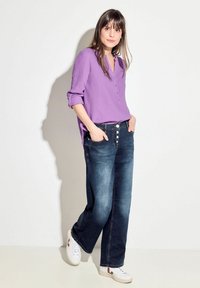 Camisa morada y texturizada con cuello en V y tres botones, combinada con unos jeans de mezclilla de pierna ancha en azul oscuro y zapatillas blancas con detalles rojos.