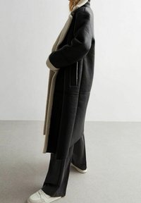 Person bærer en lang sort shearling frakke med beige foring, sorte brede bukser og hvide sneakers, stående mod en ensfarvet hvid væg.
