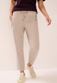 Pantalons beige à cordon de serrage avec poches latérales zippées, jambes fuselées et texture lisse. Associés à des chaussures de couleur claire.