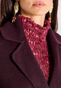 Vrouw met grote gouden en witte oorbellen, een textured rode gebreide trui en een donkerpaarse wollen jas met een brede kraag.