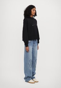 Black cropped hoodie with front logo, paired with light blue oversized jeans featuring a subtle pattern. Model wears cream sneakers.