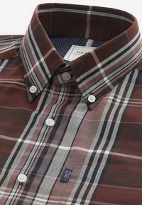Camisa de cuadros marrón con rayas blancas y azul marino, con cuello de botonadura, botones blancos y una pequeña etiqueta de logo en el frente.