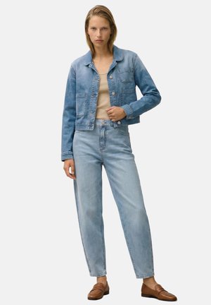 Frau steht frontal, trägt eine hellblaue Jeansjacke und Jeans, ein beiges Oberteil und braune Loafer vor einem schlichten, hellen Hintergrund.