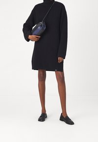 Robe-pull noire en tricot avec un col haut, des manches longues et des fentes latérales. Associée à un sac bandoulière noir et marine et des chaussures slip-on noires.