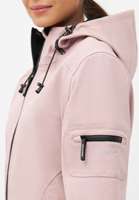 Chaqueta softshell de color rosa claro con capucha negra, cremallera frontal, cordones ajustables y un bolsillo con cremallera en la manga. Textura suave, diseño moderno.
