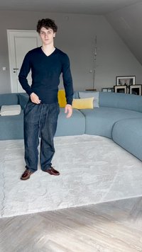 Navy v-neck sweater and loose dark denim trousers with an elastic waistband paired with brown lace-up shoes, standing on a light plush rug.