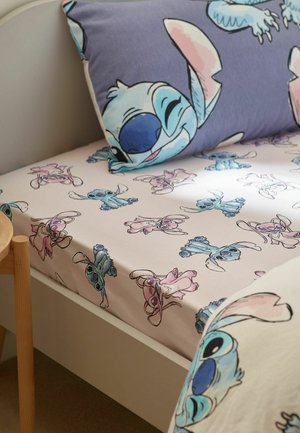 Bett mit weißem Laken und Kissen mit bunten Cartoon-Figuren in Blau und Pink, inklusive großer lächelnder Gesichter und verspielter Posen.
