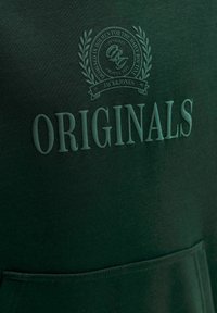 Tissu vert foncé avec texte embossé "ORIGINALS" et un emblème circulaire au-dessus, comportant des branches de laurier et un petit texte supplémentaire.
