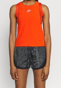 Damska koszulka na ramiączkach Nike Air w intensywnym pomarańczowym kolorze, z gładkiego materiału, bez rękawów i z siateczkowym panelem. W połączeniu z czarnymi szortami o wzorze.