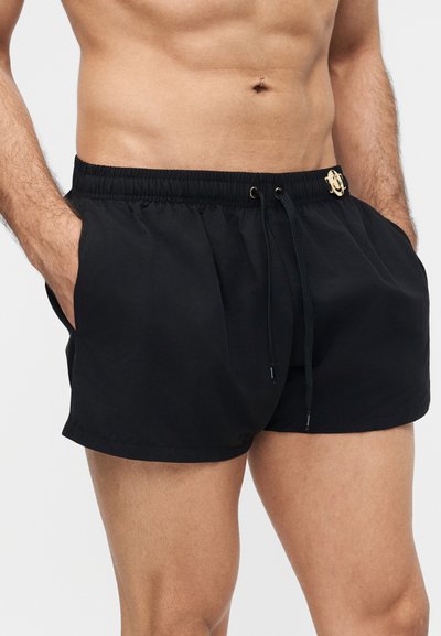 Αρσενικός κορμός που φοράει μαύρο μαγιό με ελαστική μέση, κορδόνι ρύθμισης και χρυσή λεπτομέρεια λογότυπου, με τα χέρια στις τσέπες.