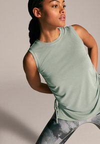 Haut de sport sans manches vert clair avec un col rond et des détails froncés sur les côtés, associé à des leggings à motifs dans des tons de gris et de noir.