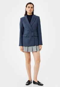 Frau steht da und trägt ein blaues, kariertes, doppelt geknöpftes Blazer, ein marineblaues Oberteil, einen hellgrauen, plissierten Minirock, silberne Creolen und schwarze Slipper.