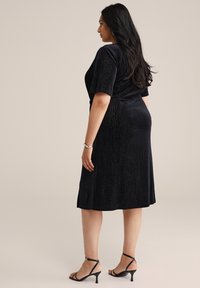Rochie neagră până la genunchi, cu mâneci scurte, realizată dintr-un material texturat, cu un luciu subtil. Pur și simplu cu sandale negre cu barete.