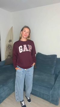 Μπορντό φούτερ GAP με λευκά και ροζ γράμματα, σε συνδυασμό με ανοιχτό μπλε φαρδιά τζιν παντελόνια. Μαύρα αθλητικά παπούτσια με λευκές σόλες, που στέκονται σε έναν καναπέ.