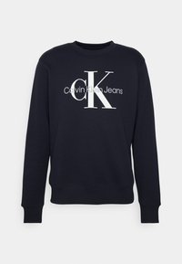 Granatowy długi sweter z białym logo "CK" i szarym logo "Calvin Klein Jeans" umieszczonym na środku klatki piersiowej.
