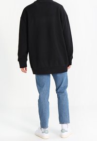 Černá oversized mikina s pokleslými rameny, žebrovanými manžetami a spodním lemem, zkombinovaná se světlými modrými džíny a bílými botami, z pohledu zezadu.