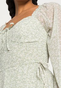 Kukkamekko vaaleanvihreänä, jossa on istuva vartalo, puffi hihat ja solmittava yksityiskohta vyötäröllä. Kangas on pehmeä ja tekstuuri on mielenkiintoinen.