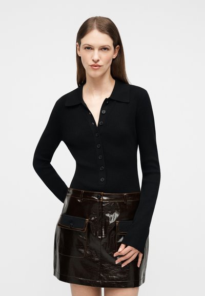 Γυναίκα με ίσια καστανά μαλλιά φορά μπλούζα μαύρη στενή με κουμπιά και μακριά μανίκια, καθώς και γυαλιστερή μίνι φούστα από σκούρο καφέ δέρμα με τσέπες στο μπροστινό μέρος.