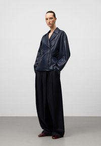 Donna in piedi con una grande giacca di pelle nera, pantaloni larghi neri e scarpe borgogna contro uno sfondo grigio uniforme.