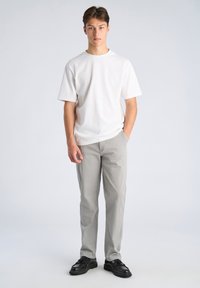 Weißes Baumwoll-T-Shirt, graue zulaufende Hosen, schwarze Schnürschuhe. Modell steht frontal mit den Händen in den Taschen vor neutralem Hintergrund.