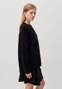 Black knitted sweater with a textured finish, round neckline, and long sleeves. Loose fit, worn over a black skirt.