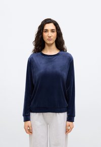 Γυναίκα με σγουρά σκουρόχρωμα μαλλιά, ντυμένη με μακρυμάνικη μπλούζα σε σκούρο μπλε και ανοιχτό γκρι παντελόνι, στέκεται μπροστά σε απλό λευκό φόντο.