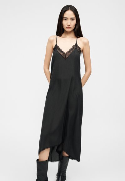 Γυναίκα με μακριά σκούρα μαλλιά φοράει μαύρο αμάνικο φόρεμα με δαντελένιο τελείωμα, συνδυασμένο με μαύρες μπότες μέχρι τη μέση της γάμπας, στέκεται μπροστά σε απλό φόντο.