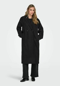 Juodas dvigubas paltas, pagamintas iš vilnos mišinio, su plačiomis atlapomis, ilgomis rankovėmis ir šoninėmis kišenėmis. Derinama su juodomis platėjančiomis kelnėmis.