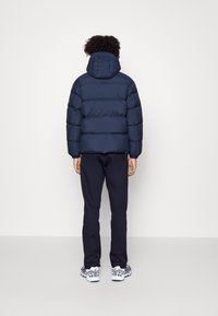 Persona con cabello rizado que lleva una chaqueta acolchada con capucha de color azul marino, pantalones oscuros y zapatillas blancas, de pie y de espaldas frente a un fondo simple.