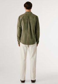 Camisa de pana verde con yugo trasero, dobladillo curvado y puños abotonados; llevada con pantalones color crema y zapatos marrones sobre un fondo neutro.