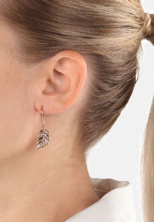 Orecchino a forma di foglia color oro con cristalli incorporati indossato all'orecchio sinistro, donna con capelli biondi raccolti, pelle chiara, colletto bianco visibile.