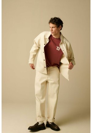 Jonge man past een open crèmekleurig jack aan over een bordeauxrood T-shirt met logo, draagt crèmekleurige broek met opgerolde boorden en donkerbruine schoenen, beige achtergrond.