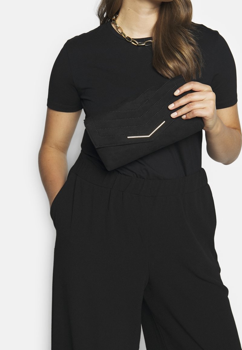 Femme portant une tenue noire et un collier en chaîne dorée tient une pochette enveloppe noire avec surpiqûres en chevron et accent doré.