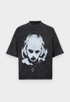 Crna oversized majica s bijelom grafikom ćelave osobe tamnih očiju, ruža, piercinga i ogrlice s križem kao privjeskom.