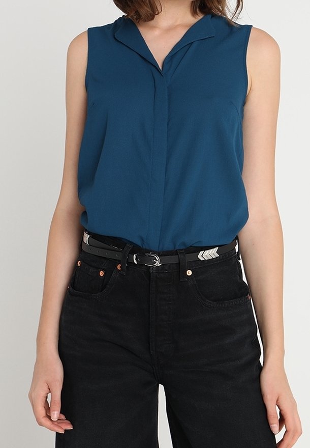 Femme portant un blouse sans manches turquoise rentré dans un jean noir taille haute avec une ceinture noire comportant des détails en chevron argentés.