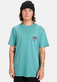 Mladý člověk s kudrnatými vlasy, který nosí černou čepici a tyrkysové triko Quiksilver s grafikou a tetováním na obou rukou.