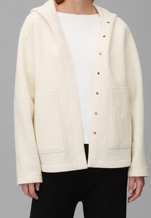 Person wearing an off-white hooded jacket with snap buttons and large front pockets over a white top and black pants.